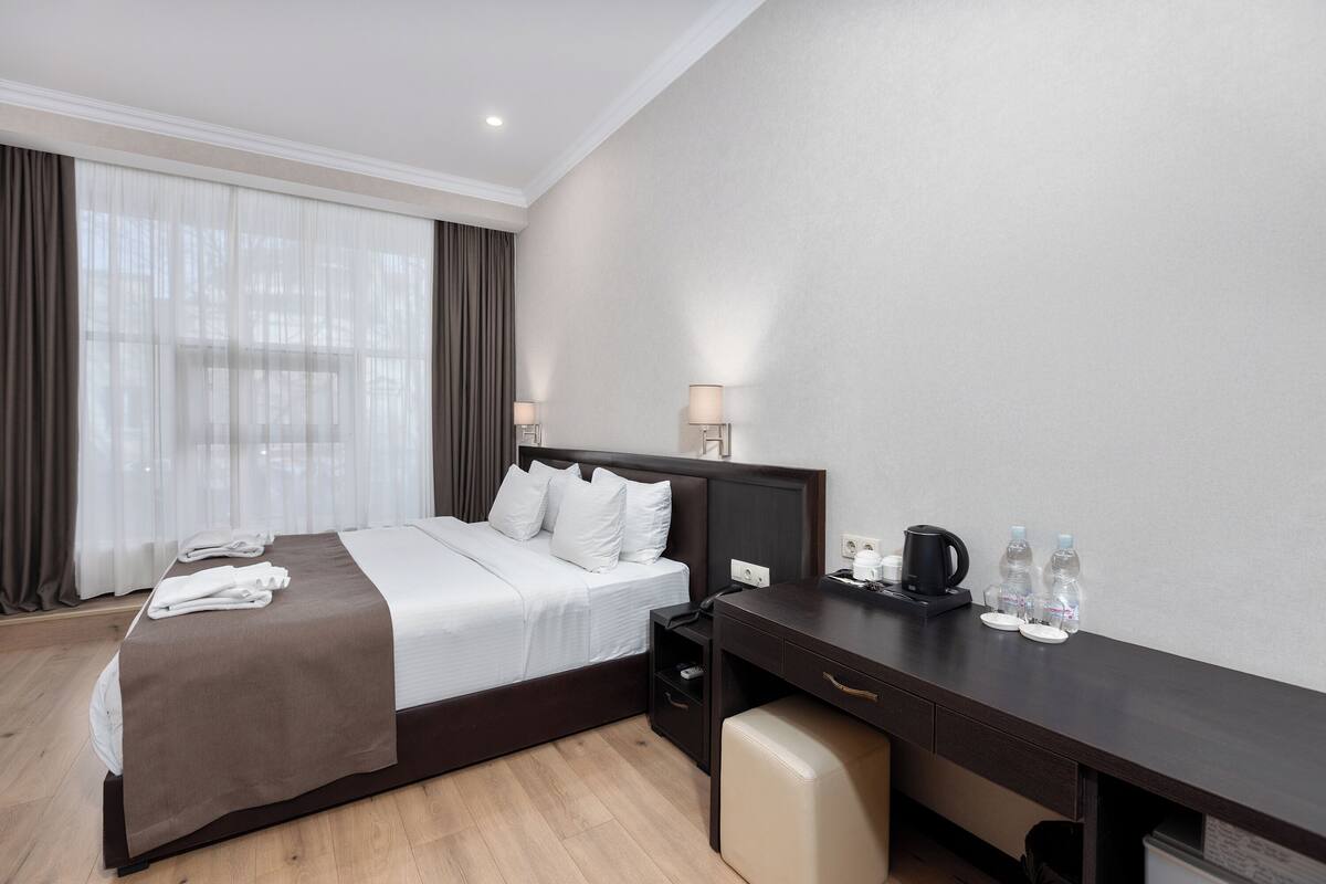 A hotel room with a bed, bedside table, desk, and a window with curtains.