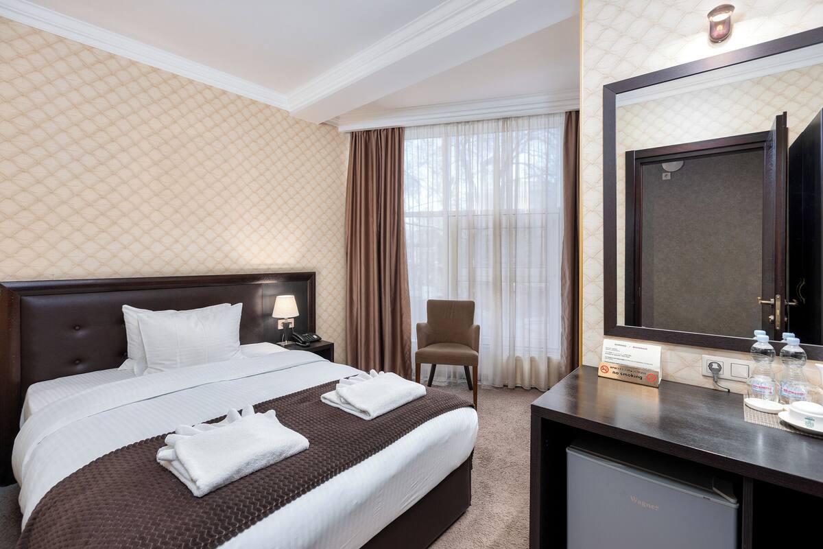 A hotel room with a bed, a desk, a chair, a mirror, and a no-smoking sign.