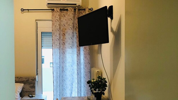Ein gemütliches Zimmer mit einem Bett, einem kleinen Tisch mit einer Pflanze, einem an der Wand befestigten Fernseher und einem Vorhang.