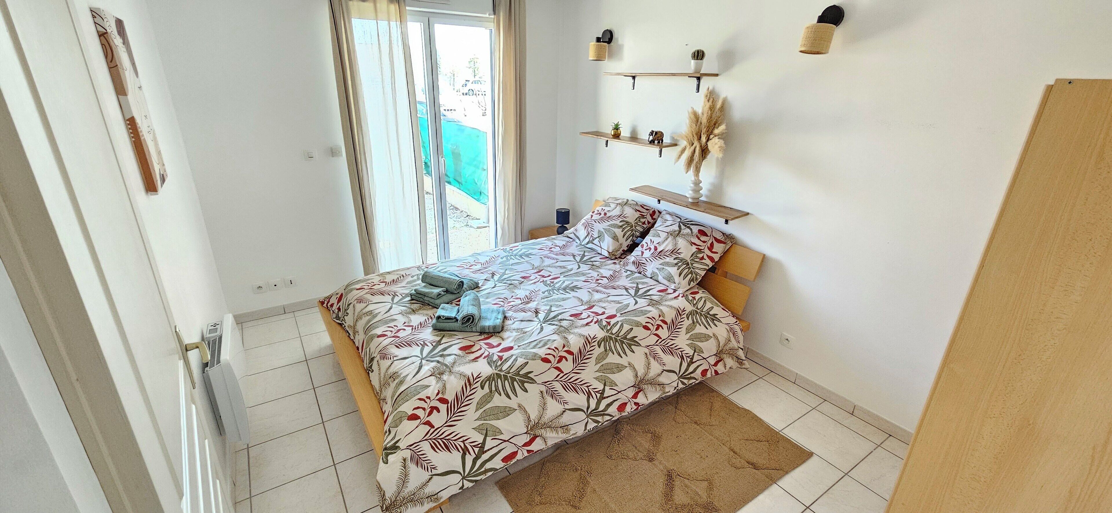 A bedroom with a bed, a window with curtains, a wall-mounted shelf with decorative items, and a door.