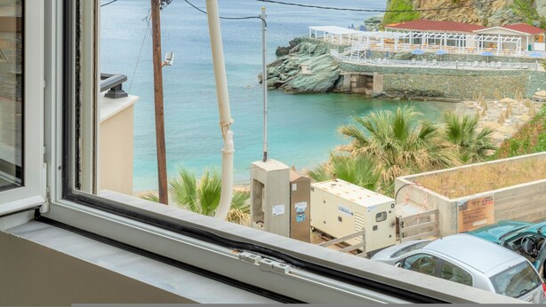 Vista costera desde una ventana con vista a una playa, un muelle rocoso y un edificio de techo rojo.