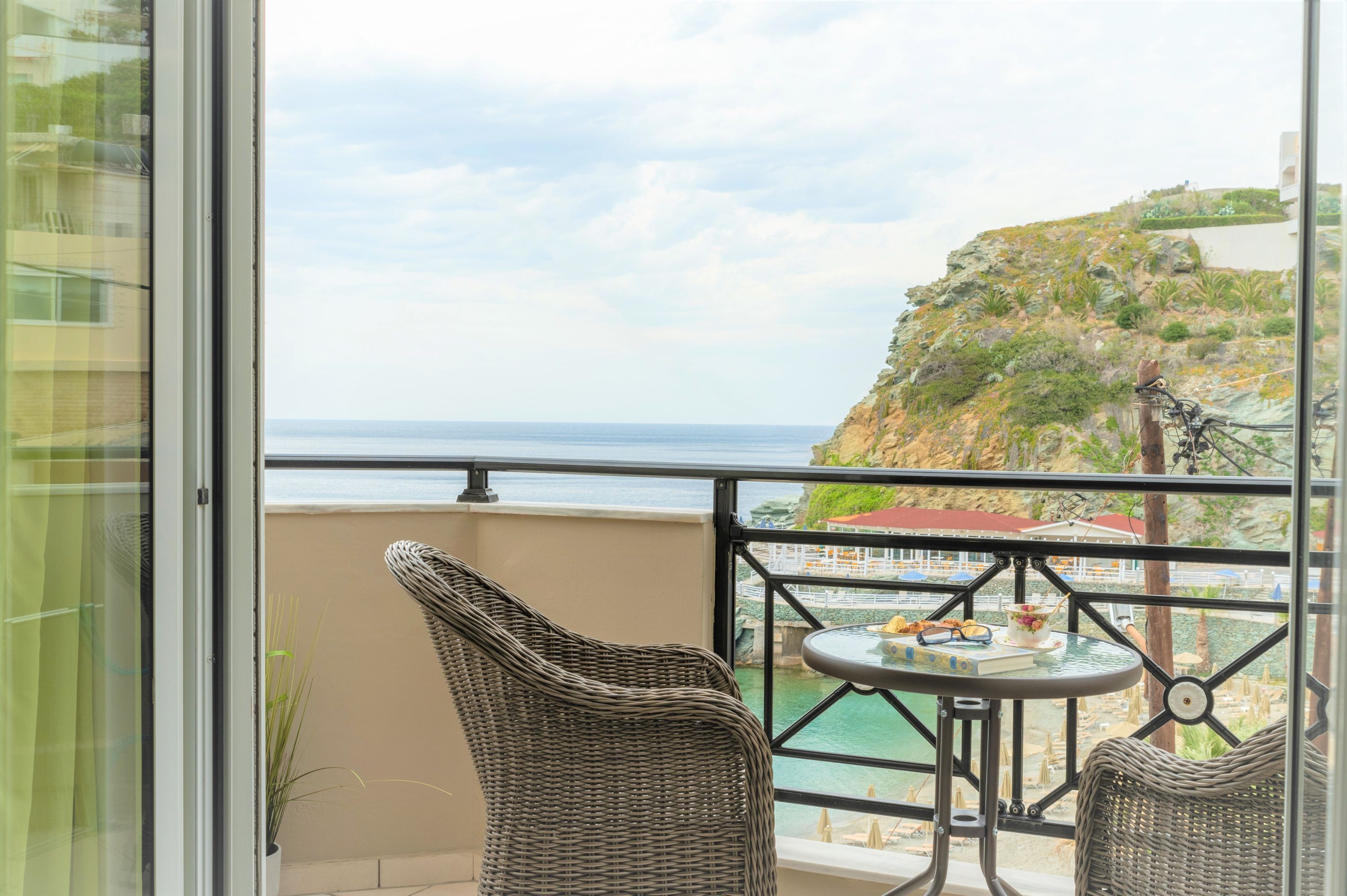 Un balcon agrémenté de fauteuils en osier, d’une petite table et offrant une vue sur la mer et une colline rocheuse.