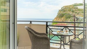 Un balcone con sedie in vimini, un tavolino e vista sul mare e su una collinetta rocciosa.