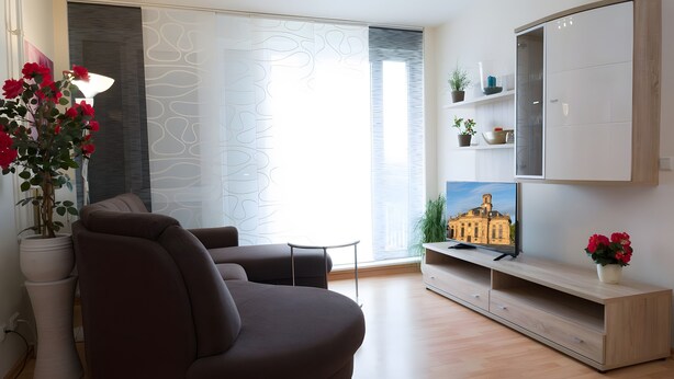 Een moderne woonkamer met een bank, een televisie op een houten dressoir, een kamerplant en een raam met uitzicht op een gebouw.