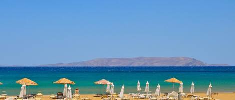 A beach with sun loungers and umbrellas, a clear sky, and distant mountains.