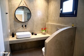 Une salle de bain avec un miroir rond, un lavabo blanc, un petit vase avec des fleurs et une fenêtre donnant sur l’extérieur.
