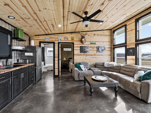 A cabin interior with a kitchen, dining area, and living room, featuring wooden walls, a ceiling fan, and large windows.