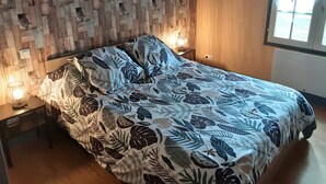 A bed with a leaf-patterned bedspread, a wooden wall panel, a bedside table with a lamp, and a window with curtains.
