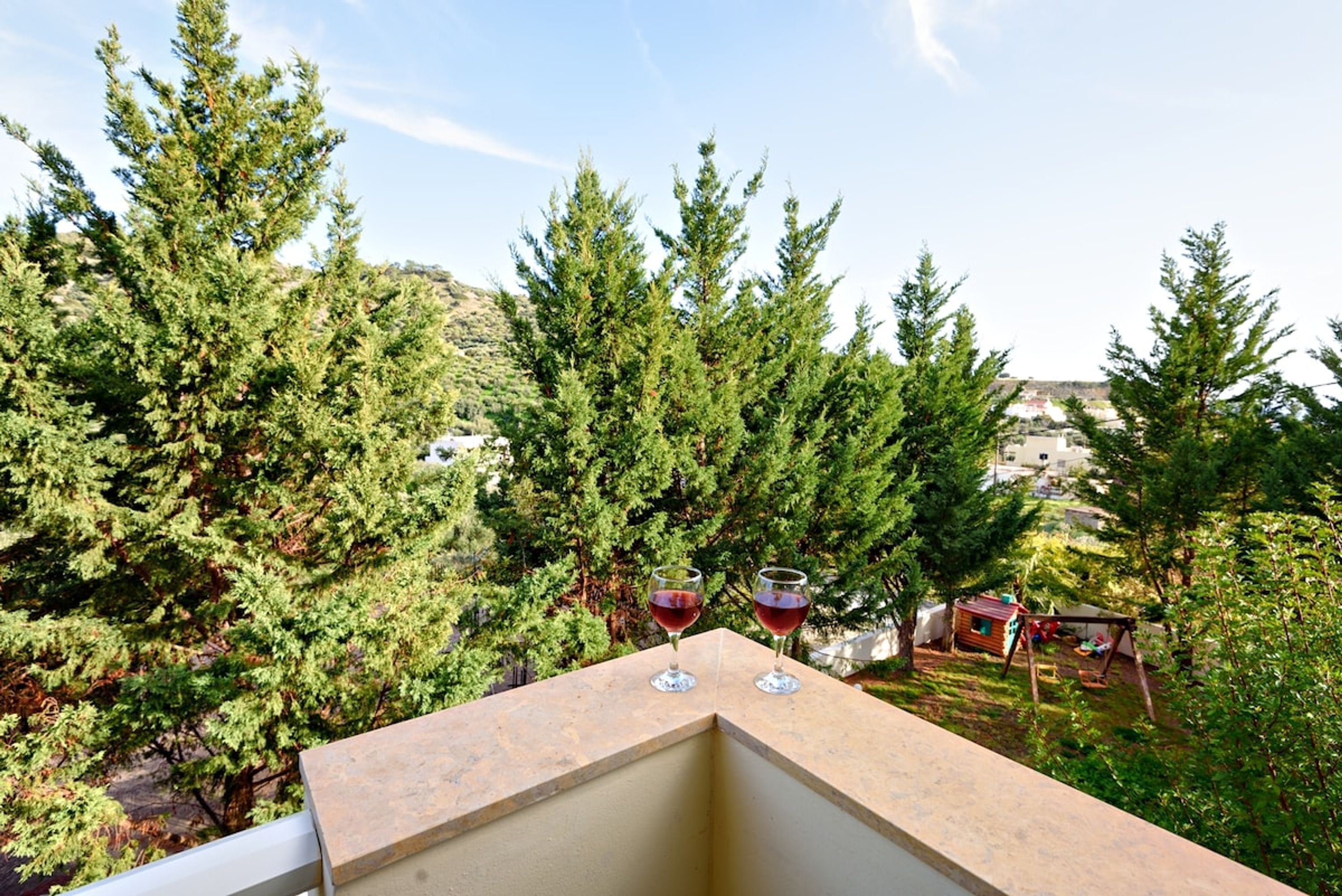A balcony with two glasses of red wine, surrounded by coniferous trees and a view of a residential area.