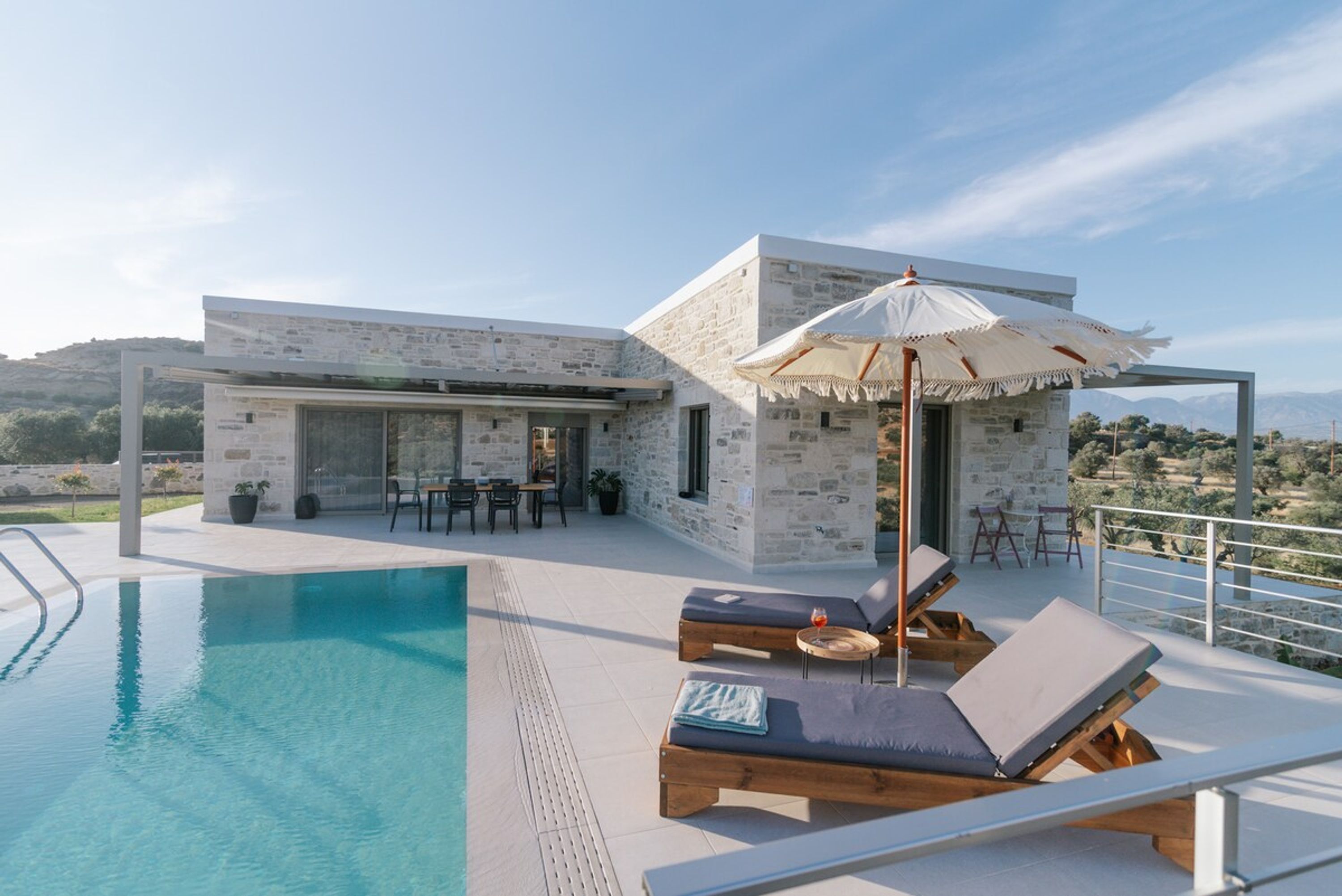 A modern pool area with lounge chairs and an umbrella, a stone building with a terrace, and a clear view of the landscape.