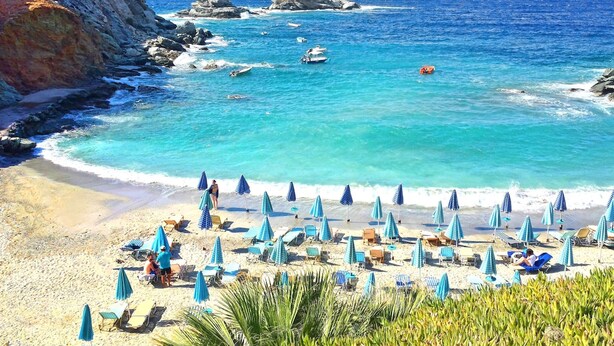 Ein Strand mit klarem, blauem Wasser, felsigen Klippen und Sonnenschirmen.