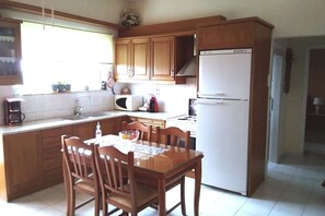 A kitchen with wooden cabinets, a white refrigerator, a dining table with chairs, and a window providing natural light. - Marina's traditional house at Sivas village (Sivas)