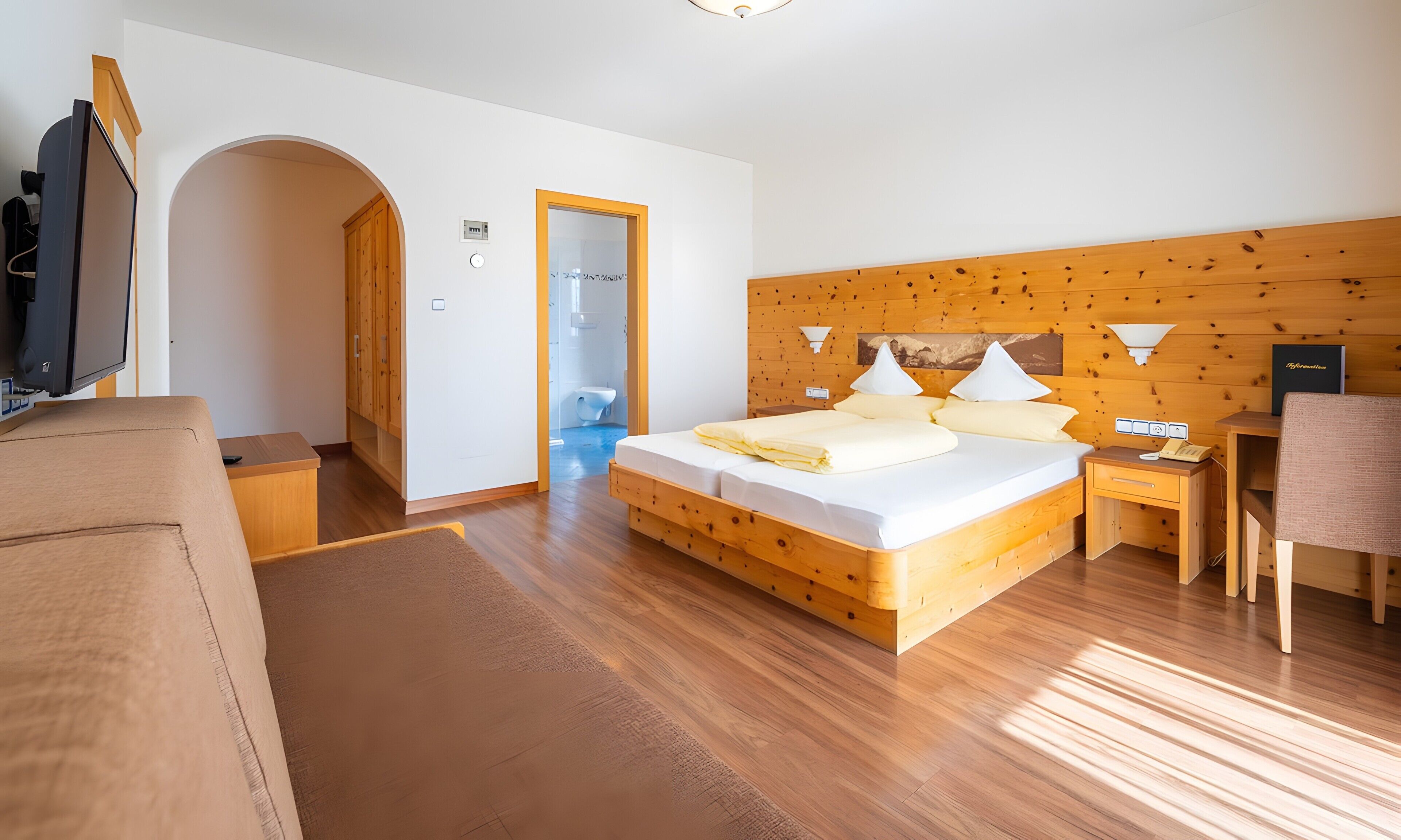 A hotel room with a wooden headboard, a bed with white bedding, a desk with a chair, a television, and a bathroom visible through an archway.