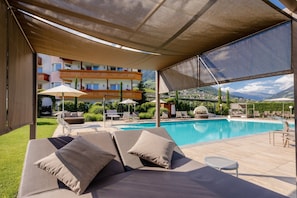 Un área junto a la piscina con sillas de descanso, una zona de estar cubierta y vistas a un edificio de varios pisos con balcones.