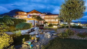 Um hotel com vários andares, piscina, jardins bem cuidados e montanhas ao fundo.