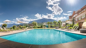 Uma piscina retangular com espreguiçadeiras e guarda-sóis, cercada por uma paisagem montanhosa e um edifício com várias andares e varandas.