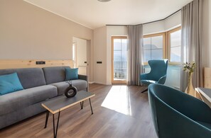 A modern living room with a sofa, a coffee table, and a chair. There is a large window with curtains, a door, and a view of the outdoors. - Room 'Passerblick Suite 4' with Mountain View, Wi-Fi and Air Conditioning (Tirolo)