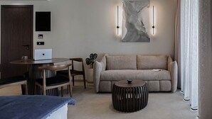 Habitación de hotel moderna con sofá, una mesita redonda de madera, un ordenador portátil sobre una pequeña mesa, una planta en maceta y un cuadro abstracto grande en la pared.