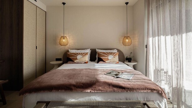 A bedroom with a large bed, two bedside tables, a nightstand, and a window with sheer curtains.