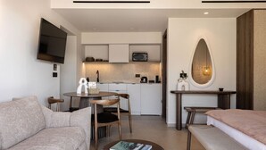 A modern hotel room with a sofa, a small round table with a book, a bench, a TV mounted on the wall, a kitchenette, and a large mirror.