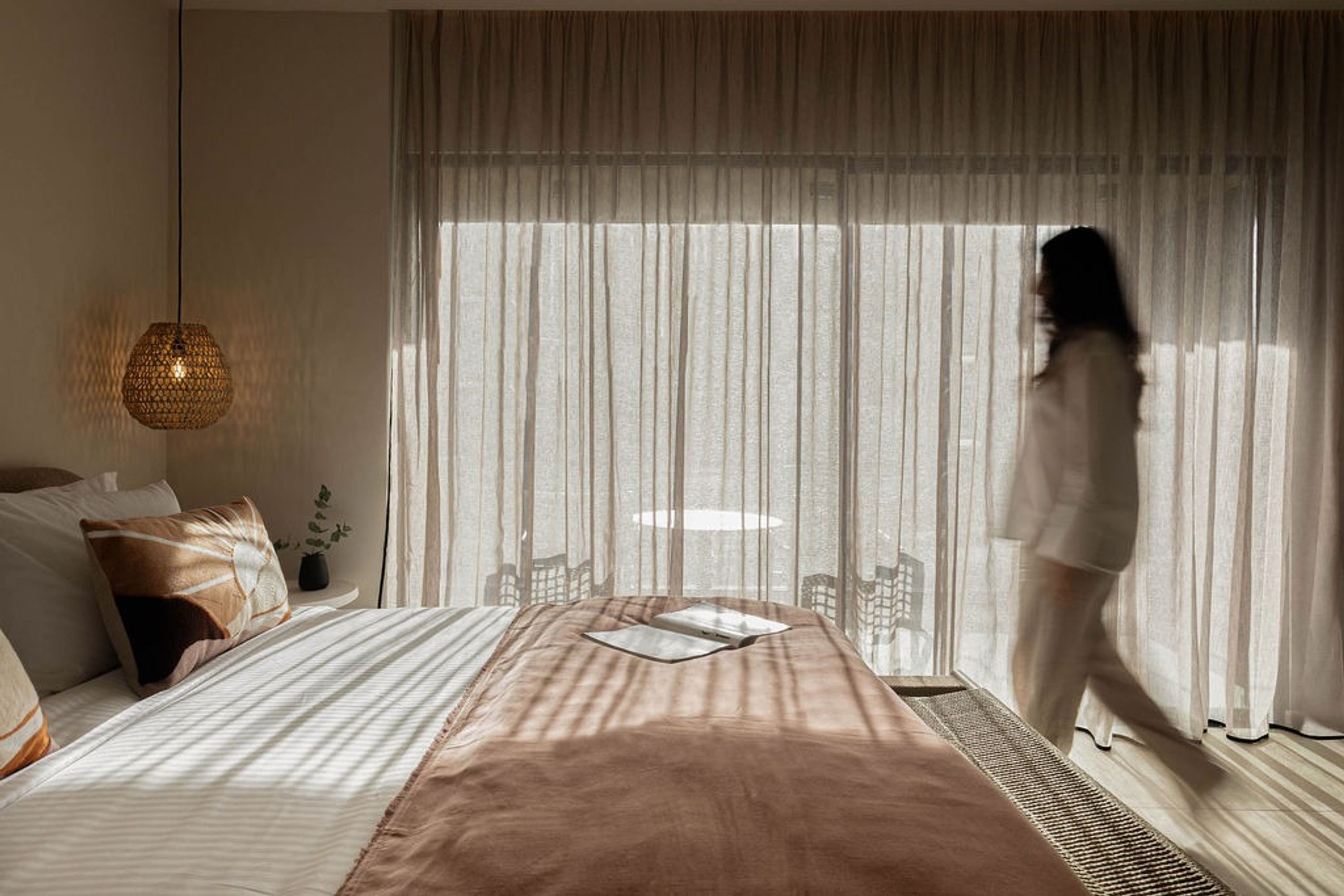 A bedroom with a large bed, a bedside table, a hanging light fixture, and sheer curtains.