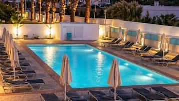 A swimming pool with lounge chairs and umbrellas, surrounded by palm trees and a view of a hill.