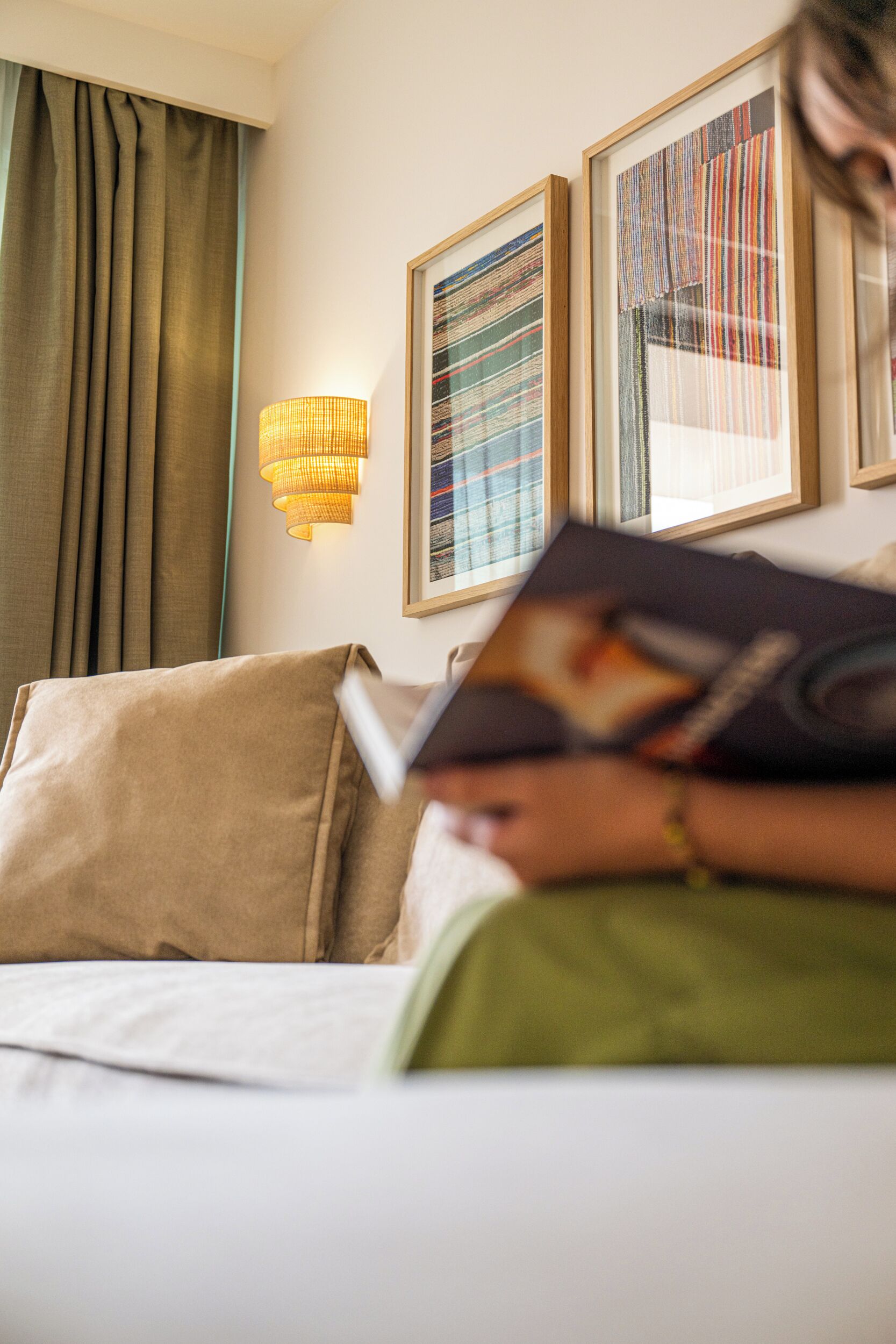 A person reading a book on a bed with a lamp and framed artwork on the wall.