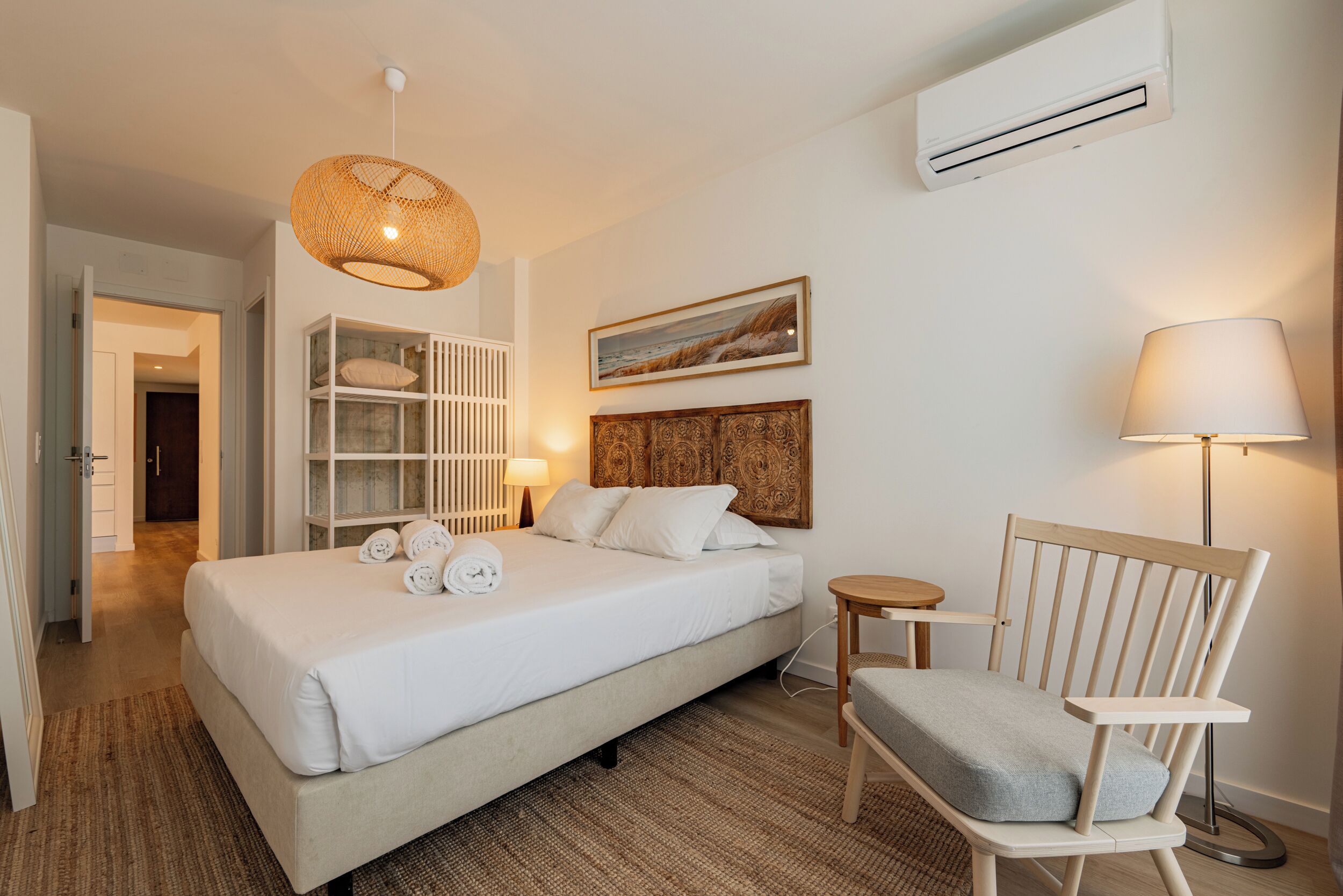 A bedroom with a bed, bedside tables, a chair, a lamp, a shelf with decorative items, and an air conditioning unit.