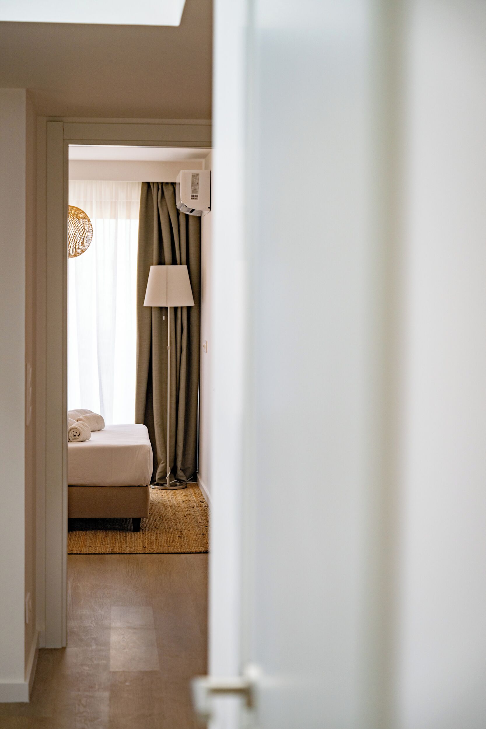 Ett hotellrum med en säng, en lampa, ett fönster med gardiner och en luftkonditioneringsanläggning.