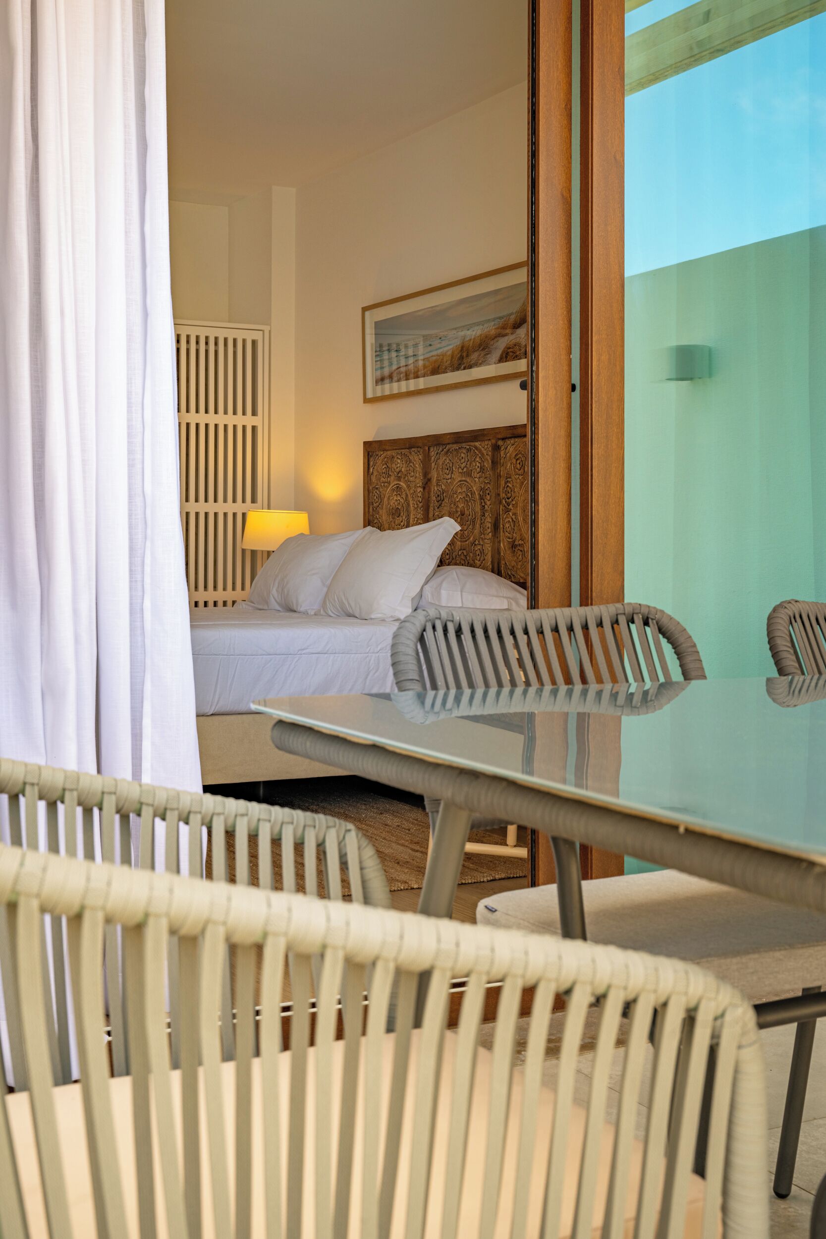 A hotel room with a bed, a glass table, wicker chairs, a lamp, and a view of the sky.