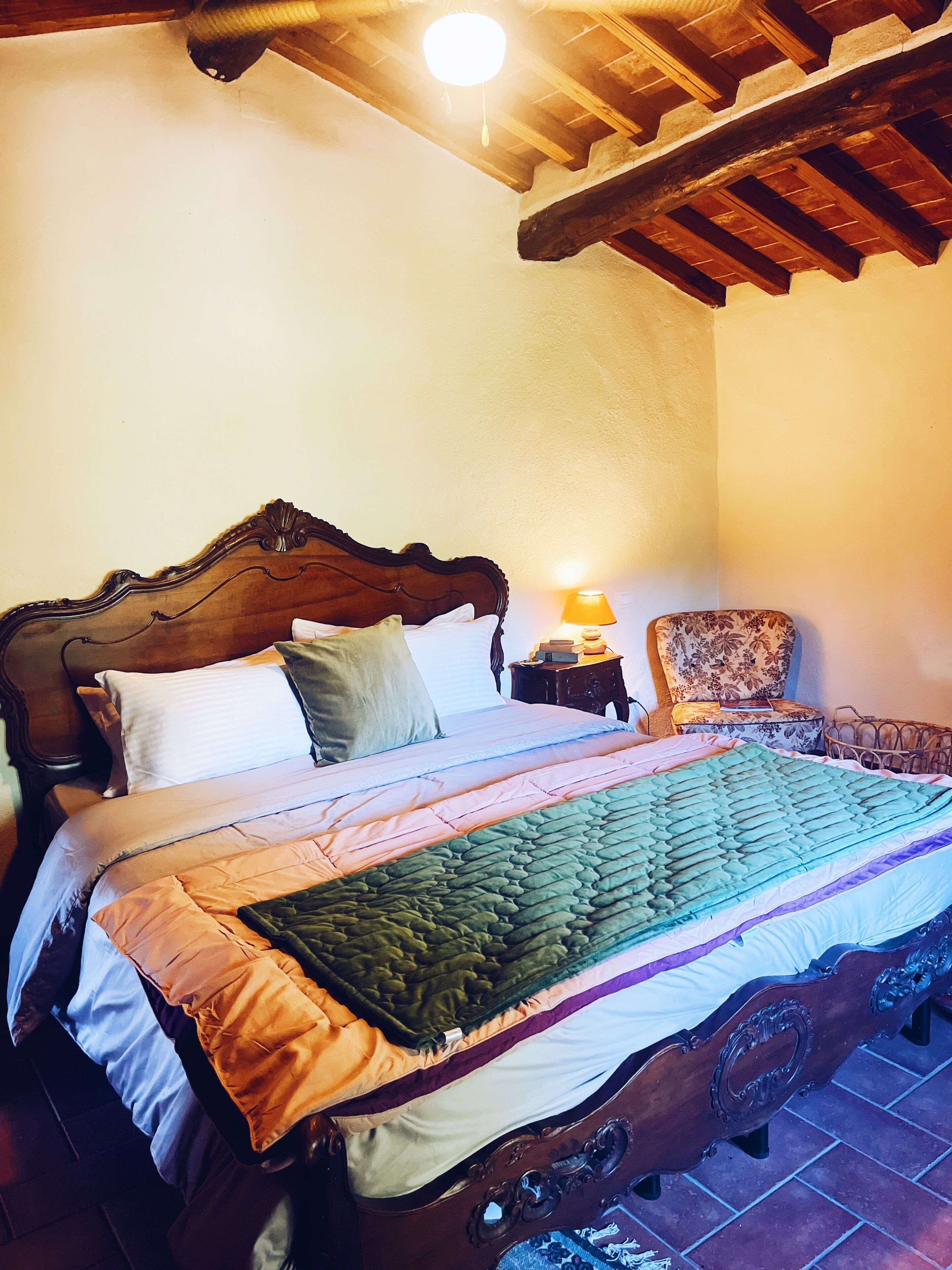 A traditional wooden bed with a headboard, a bedside table with a lamp, and a floral-patterned chair.