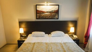 Una cama doble con una colcha estampada, dos lámparas de noche, una mesita de noche con un teléfono, y un cuadro sobre la cama.