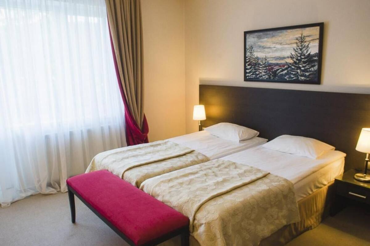 Een hotelkamer met twee bedden, een bankje, een schilderij en een raam met gordijnen.