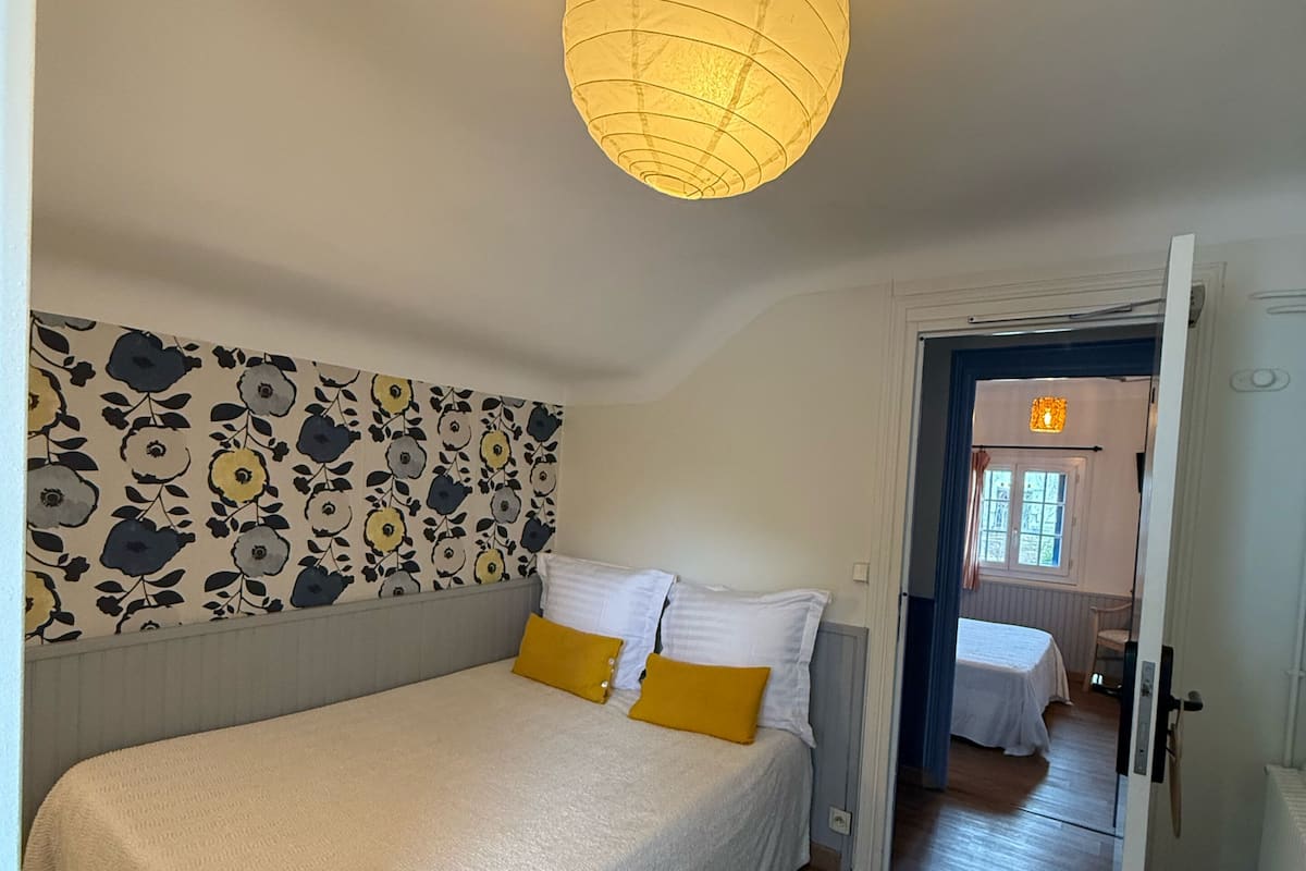 Un dormitorio con una cama, un papel tapiz estampado, una lámpara en el techo y una puerta que da a otra habitación.