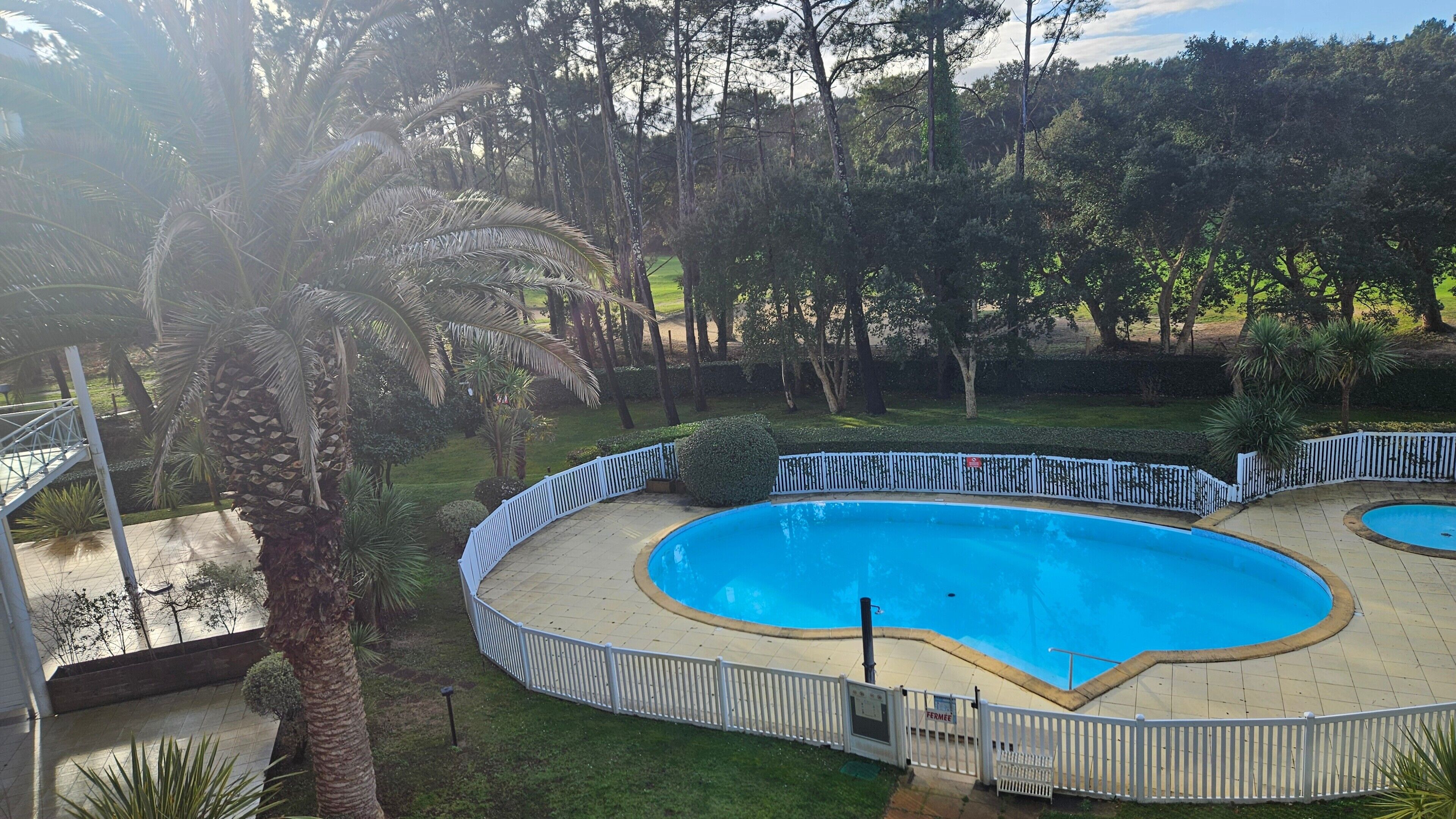 Une piscine entourée d’une clôture blanche, bordée d’arbres et d’herbe.