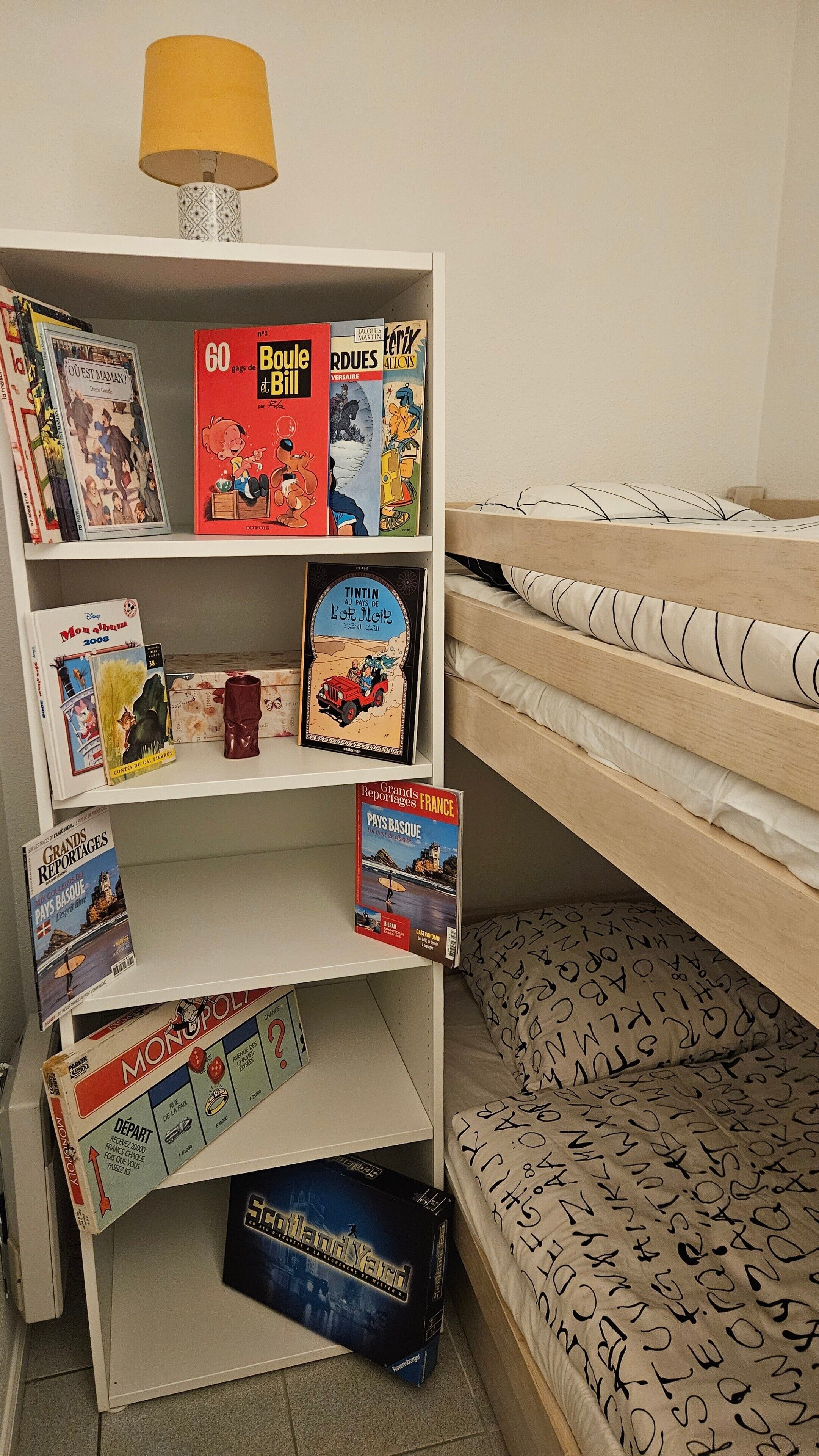 A room with a bunk bed, a bookshelf with various books and board games, and a lamp on the shelf.