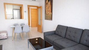 A living room with a grey sofa, a glass coffee table, a dining table with chairs, and a kitchen area with cabinets and a refrigerator. - Anacasa Albatros MET Oliva Nova Golf AP465 (Oliva)