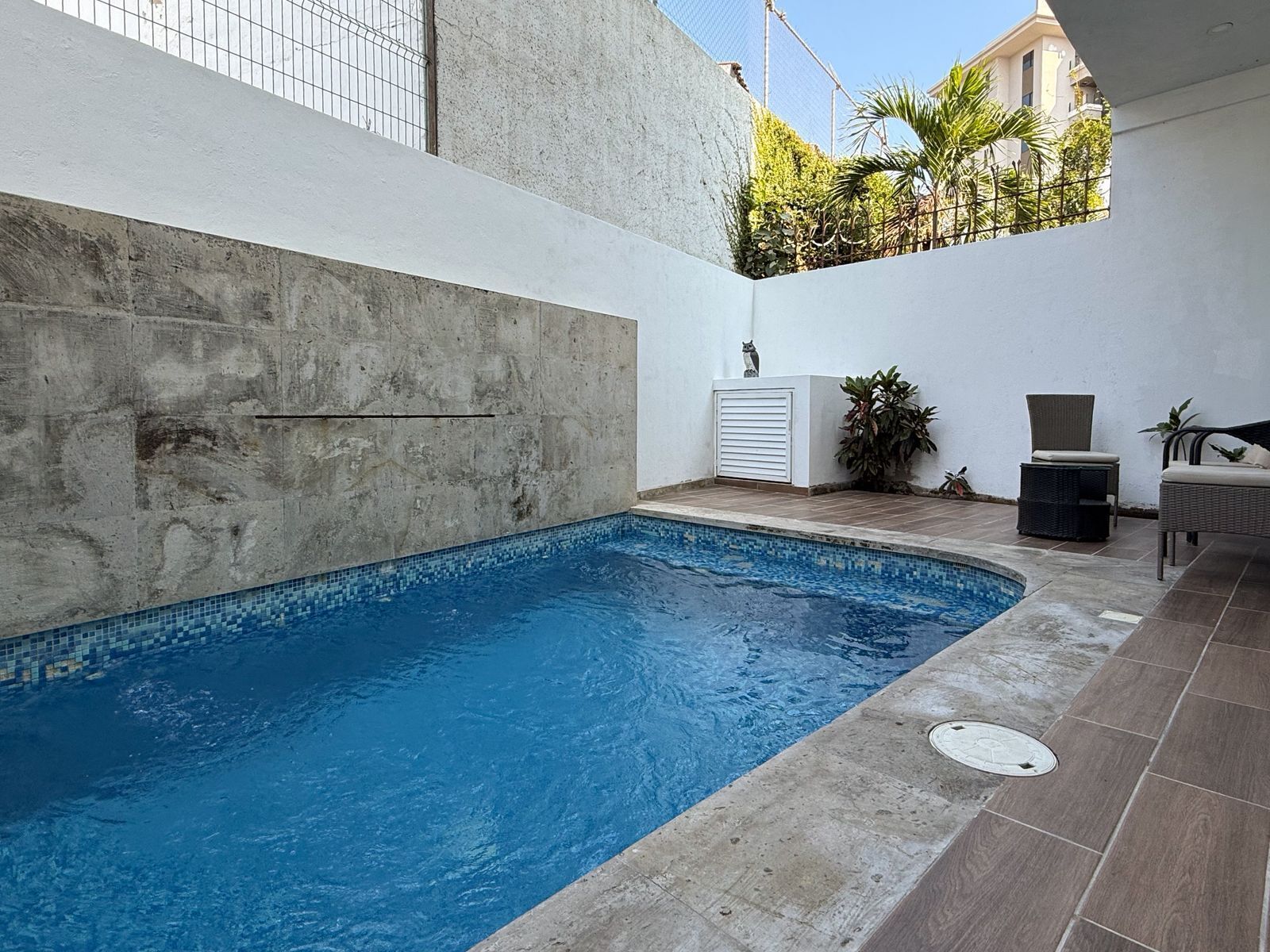 A swimming pool with tiled edges, a concrete wall, and a small outdoor seating area with chairs and a table.