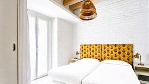 Una camera da letto con una parete in mattoni bianchi, un'ampia finestra, un letto con biancheria bianca, una testiera gialla con motivo e un lampadario.