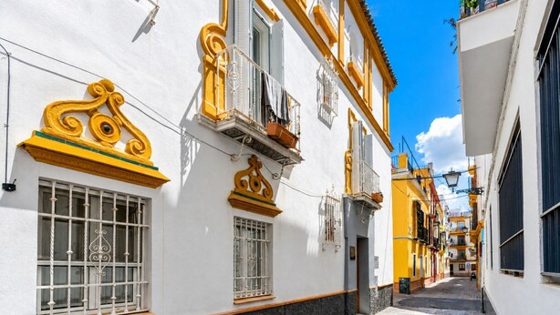 A narrow street with white buildings featuring yellow accents and ornate window designs. - Casa San Vicente Olehousing 3 (Sevilla)
