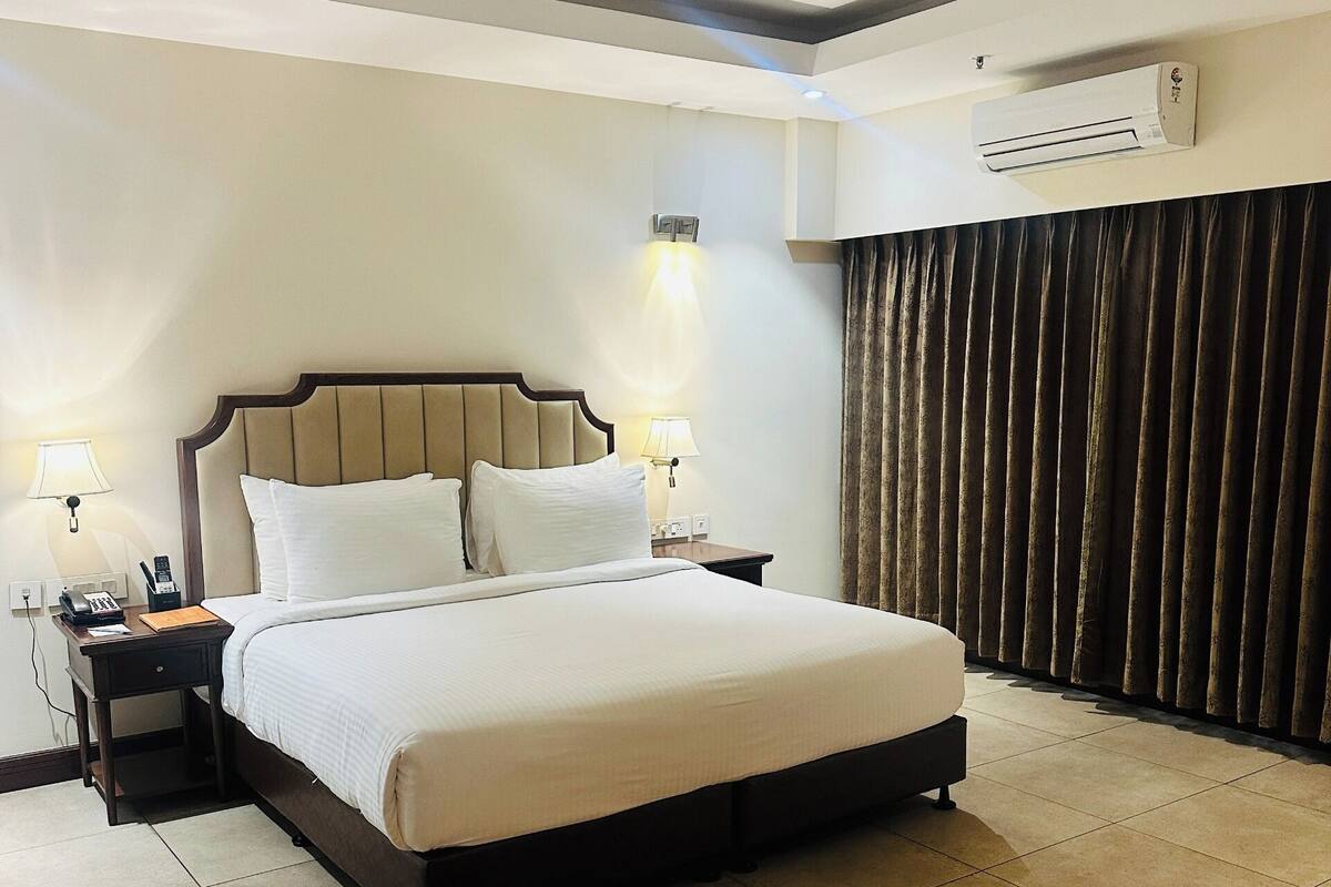 Una habitación de hotel con cama, lámparas de noche, ventilador de techo, unidad de aire acondicionado y cortina.