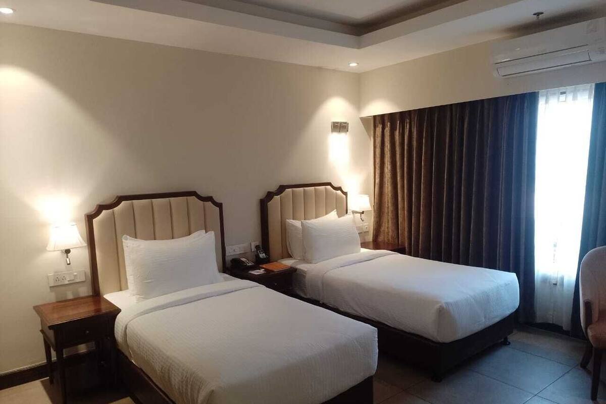 Habitación de hotel con dos camas, una mesita de noche, una lámpara, una silla y una ventana con cortinas.