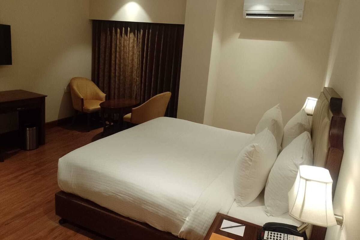 Habitación de hotel con cama, escritorio, dos sillas, lámpara, equipo de aire acondicionado y teléfono.