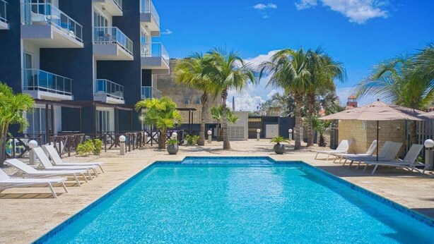 A swimming pool with lounge chairs, palm trees, and a modern building with balconies. - 3-Suite Condo Steps to Palm Beach (Noord)