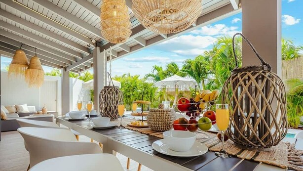 Un área de comedor al aire libre con lámparas de mimbre, una piscina y abundante vegetación de fondo.