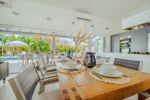 A dining area with a long wooden table set for a meal, surrounded by chairs, with a view of a pool and greenery outside. - Villa Lua (Noord)