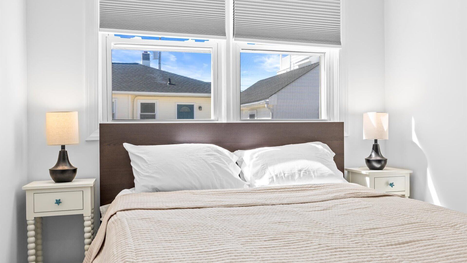 A bedroom with a bed, two bedside tables with lamps, a window with blinds, and a view of a building outside.