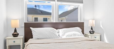 A bedroom with a bed, two bedside tables with lamps, a window with blinds, and a view of a building outside.