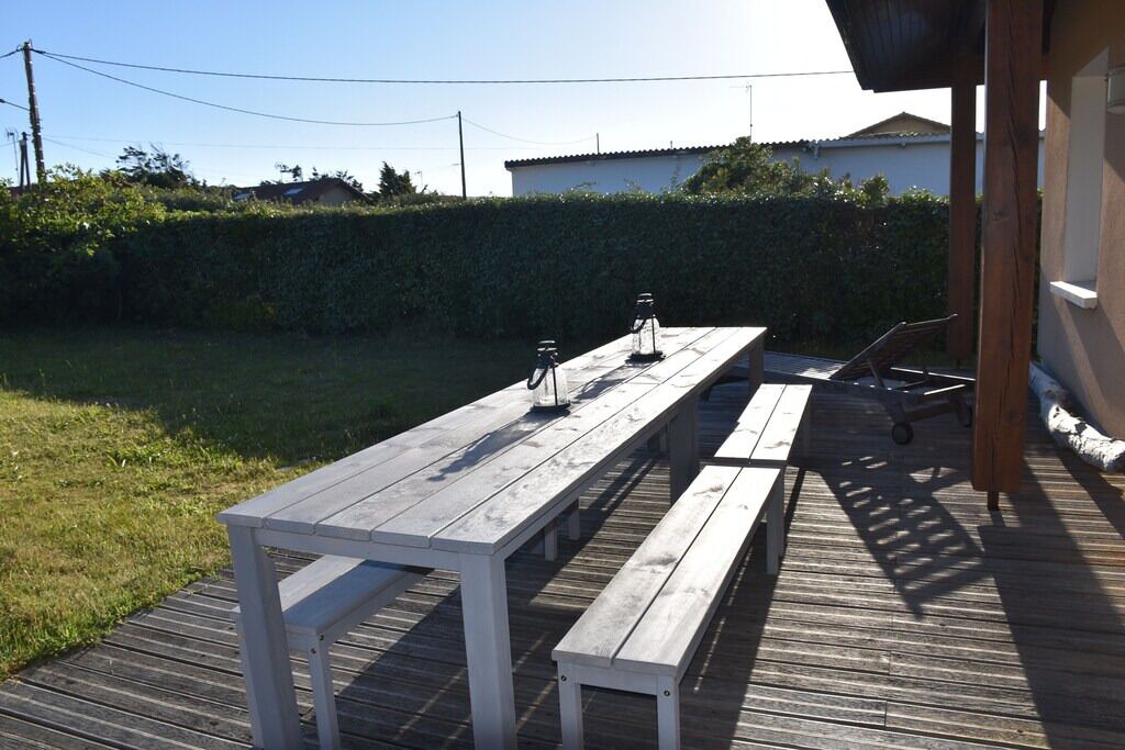 Une terrasse en bois avec une table de pique-nique et des bancs, entourée d’une haie et avec un bâtiment visible au loin.