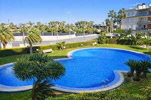 Una piscina de borde curvo, rodeada de palmeras y vegetación, con una carretera y edificios al fondo.